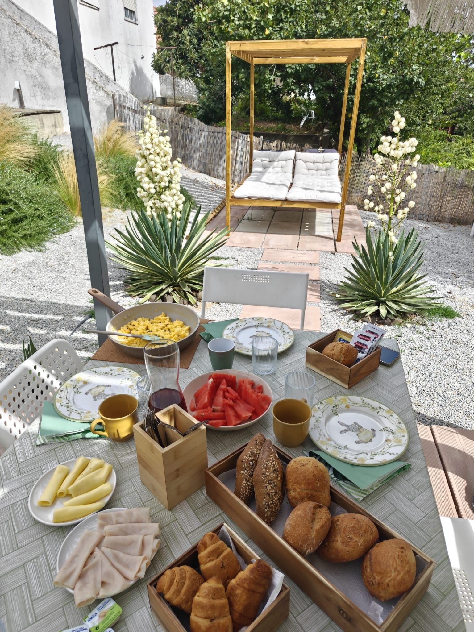 An outdoor dining area with a table set for breakfast, including bread, pastries, and fruit.