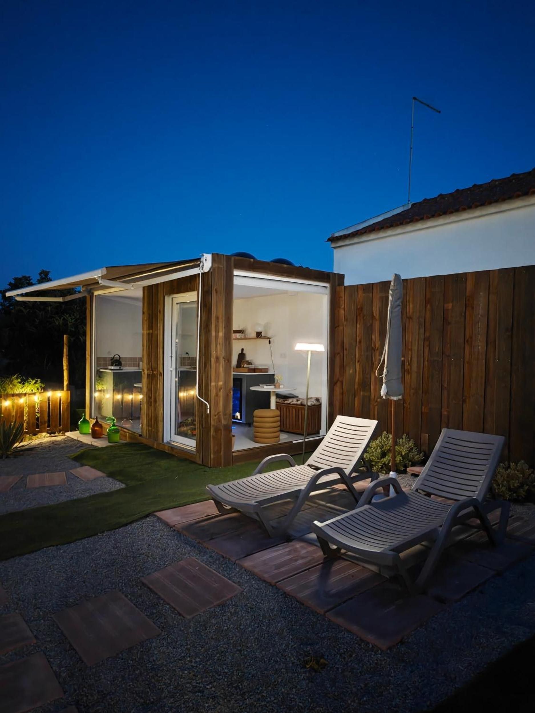 A modern outdoor patio area with a wooden deck, two lounge chairs, and a glass-enclosed structure with a kitchen visible inside.