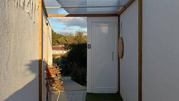 Un estrecho pasillo exterior con una puerta, un pequeño jardín con plantas y un suelo de baldosas.