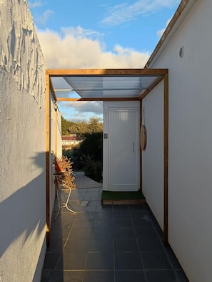 Un étroit couloir extérieur avec une porte, un petit espace jardin agrémenté de plantes et un sol carrelé.