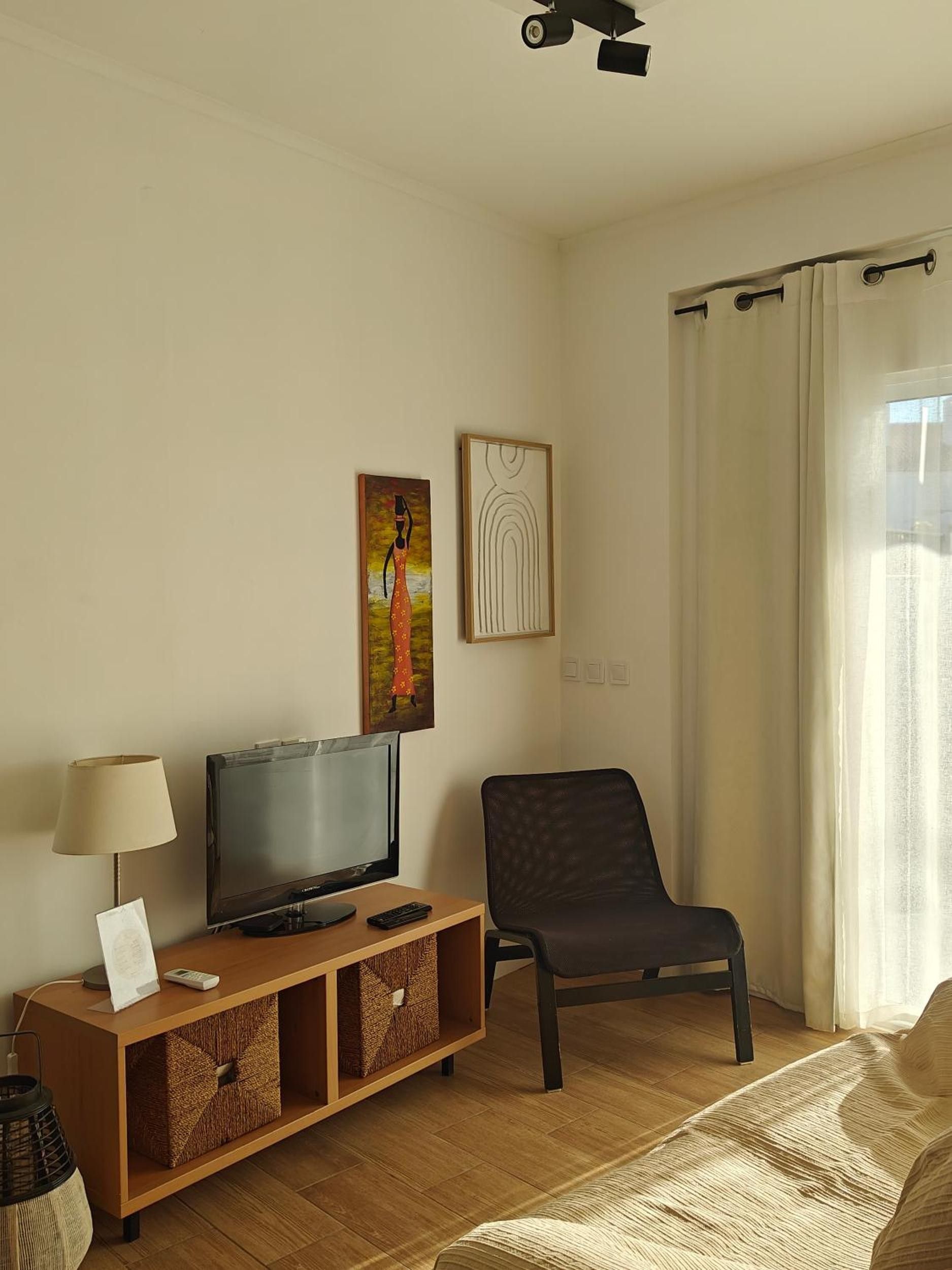 A hotel room with a television on a wooden stand, a black chair, a bed with a textured blanket, and a window with curtains.