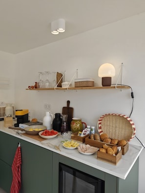 Cozinha com bancada branca, diversos alimentos, máquina de café e luminária pendente.