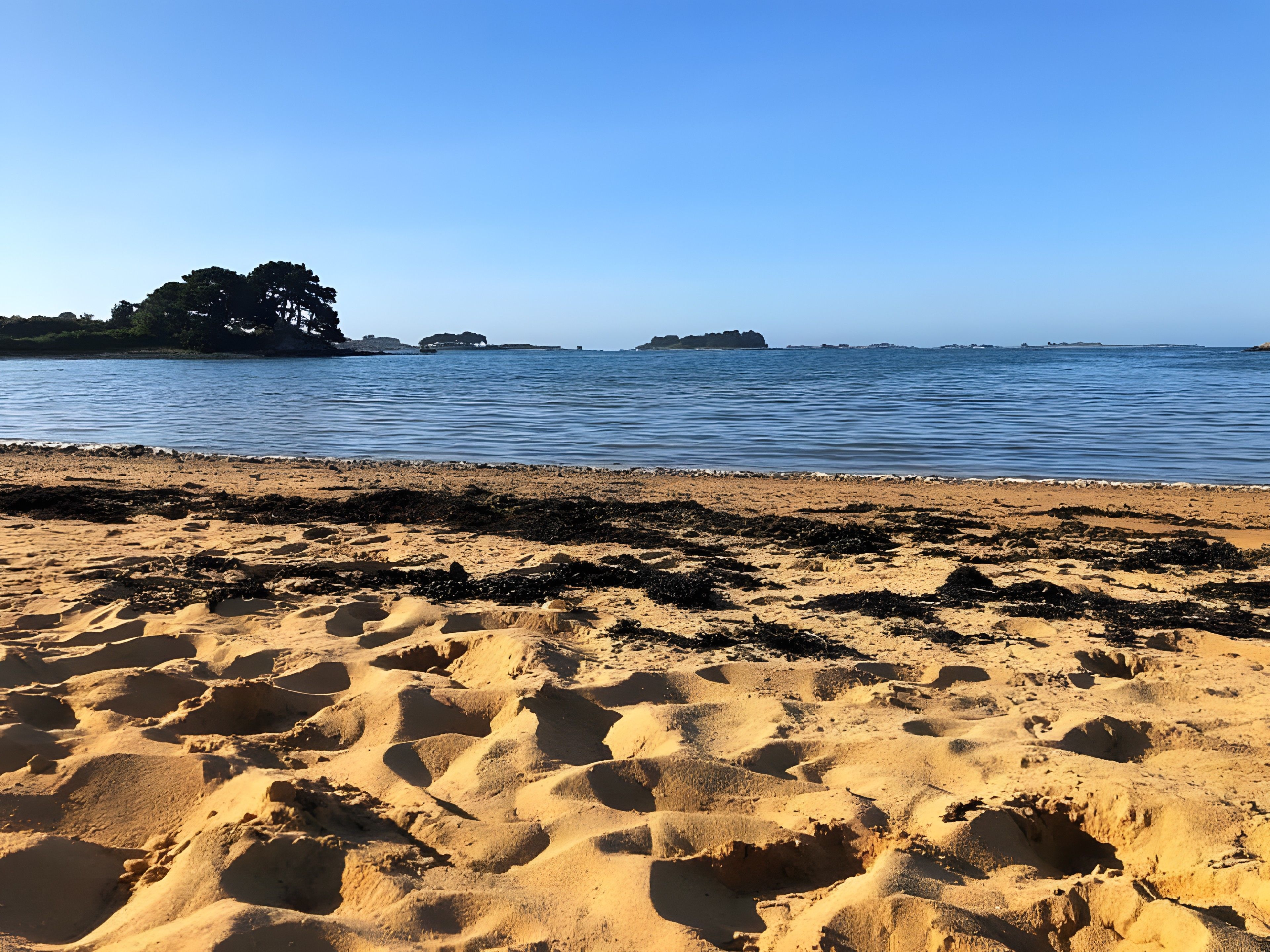 Une plage de sable où l’on peut voir des empreintes de pas menant à l’eau, un ciel dégagé et des îles lointaines.