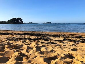 Ein sandiger Strand mit sichtbaren Fußspuren, die zum Wasser führen, ein klarer Himmel und entfernte Inseln.