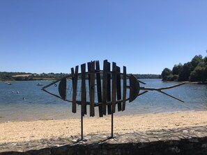 A metal sculpture with a circular base and vertical supports, located on a rocky shore with a view of a calm lake and boats in the distance.