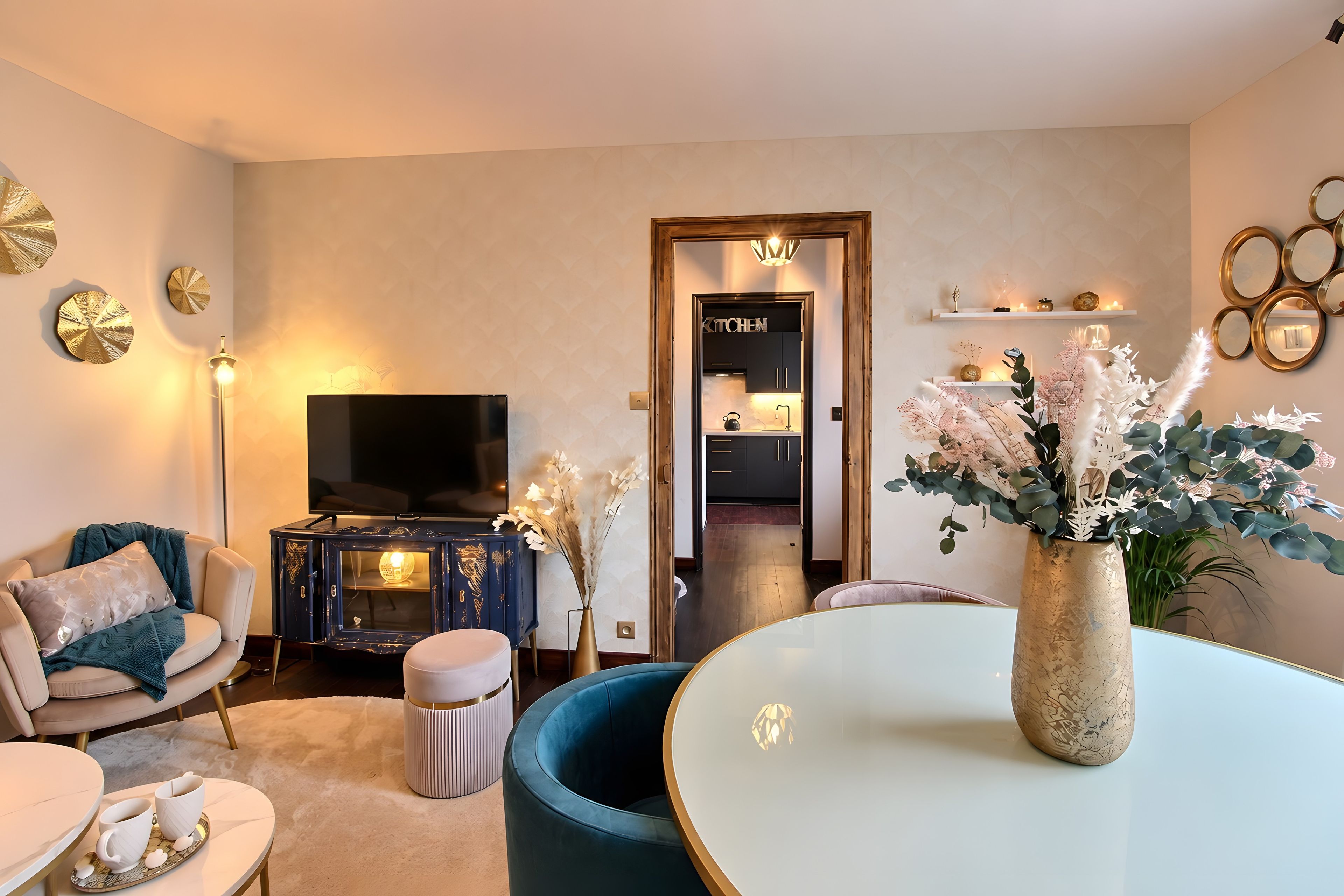 A modern living room with a television, seating, decorative items, and a kitchen area.