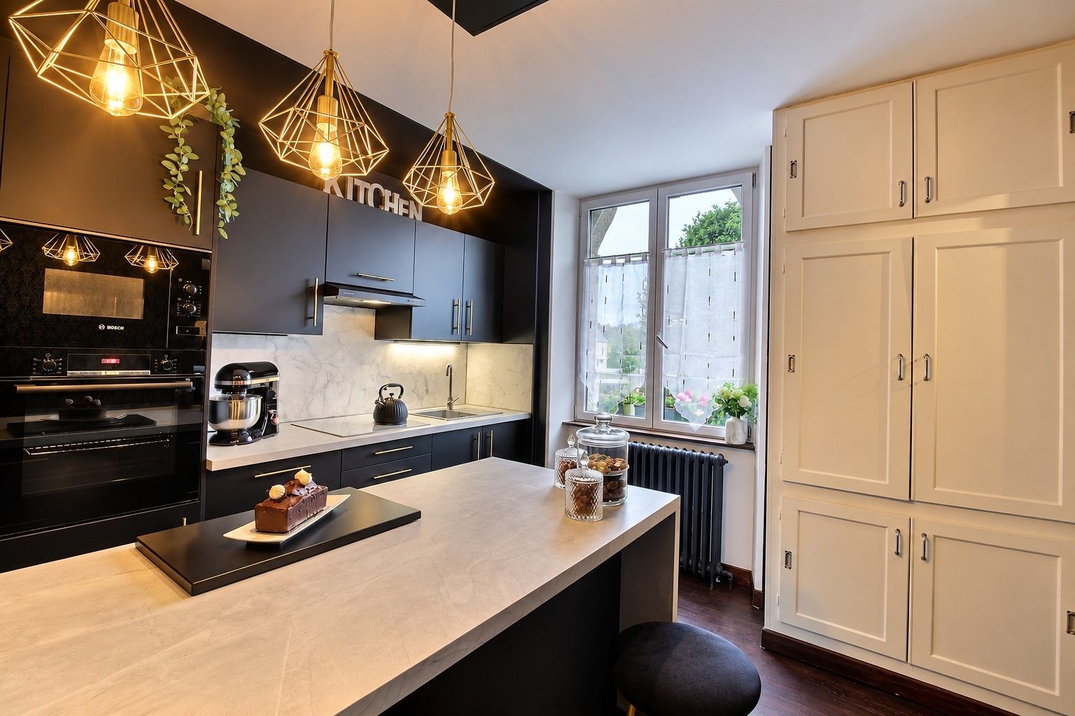 Une cuisine moderne équipée d’un four encastr é, d’une machine à café et d’une bouilloire posée sur le plan de travail. On y trouve des placards avec des poignées, une fenêtre donnant sur un paysage agréable et un radiateur.