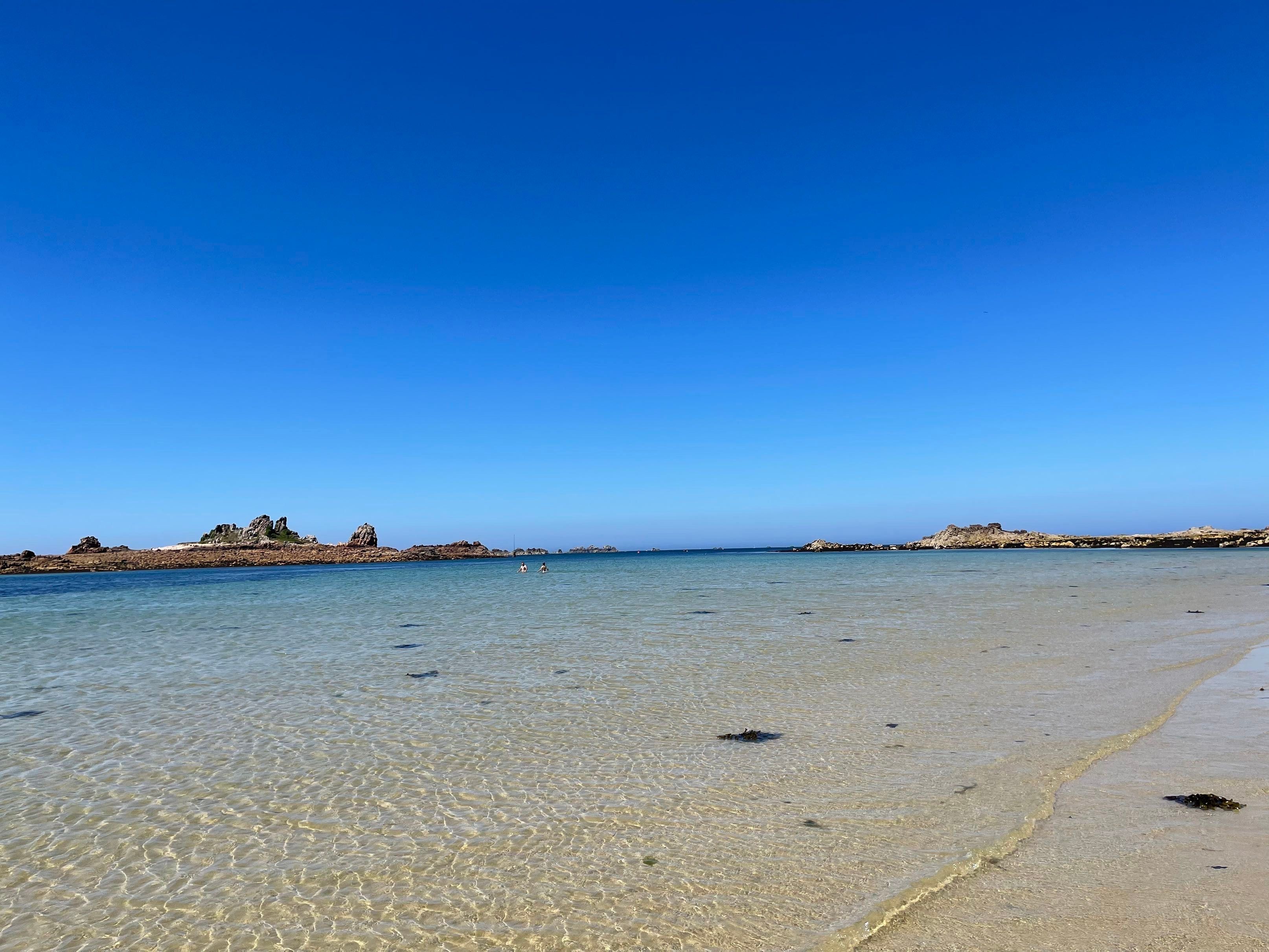 Une plage à l’eau claire, bordée d’affleurements rocheux, et un ciel dégagé.