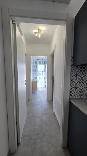En gang med en loftlampe, en dør der fører til et rum med mønstrede gardiner, og et flisegulv.