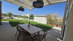 En udendørs terrasse med et bord, stole og en hængende lampe, samt udsigt over et boligområde.