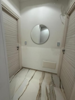 Um corredor com duas portas fechadas, um espelho redondo na parede e um chão com padrão marmorizado.