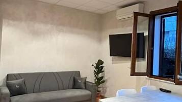 A room with a grey sofa, a white table with chairs, a TV on the wall, and a window.