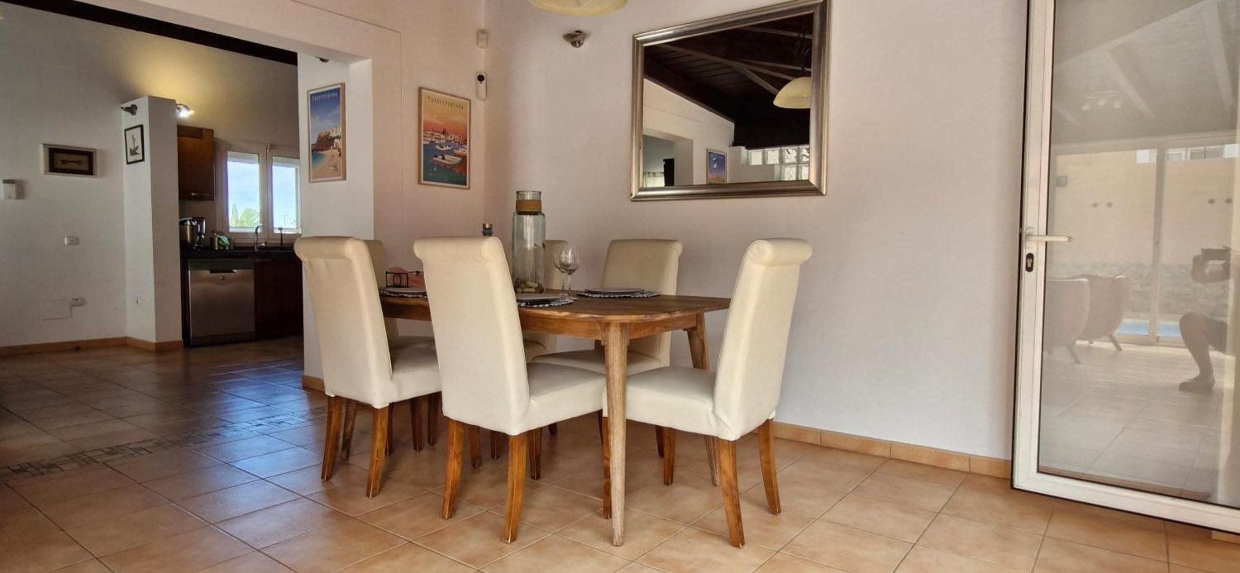Un coin repas avec une table en bois et quatre chaises, un grand miroir au mur et une cuisine à l’arrière-plan.