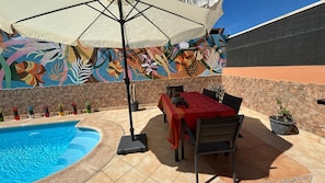 Une terrasse extérieure avec une piscine, une table entourée de chaises et une fresque colorée sur le mur.