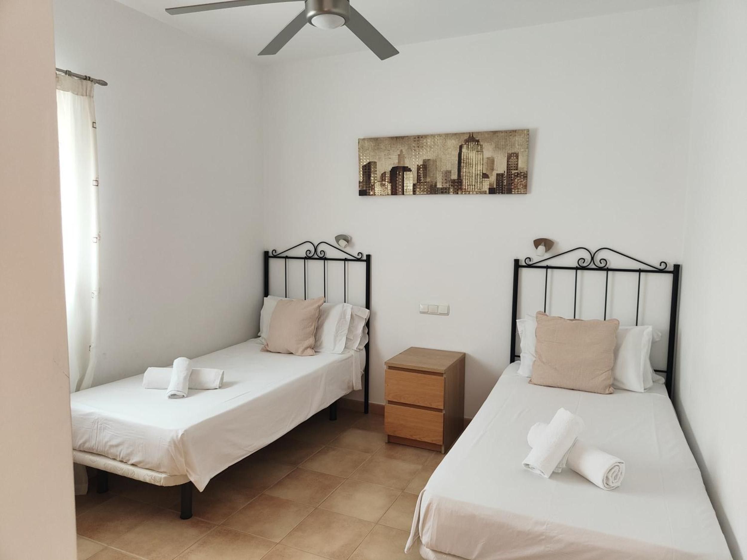 Ein Doppelzimmer mit zwei Einzelbetten, einem kleinen Nachttisch aus Holz, einem Deckenventilator und einem Stadtbild an der Wand.