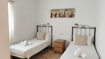 Ein Doppelzimmer mit zwei Einzelbetten, einem kleinen Nachttisch aus Holz, einem Deckenventilator und einem Stadtbild an der Wand.