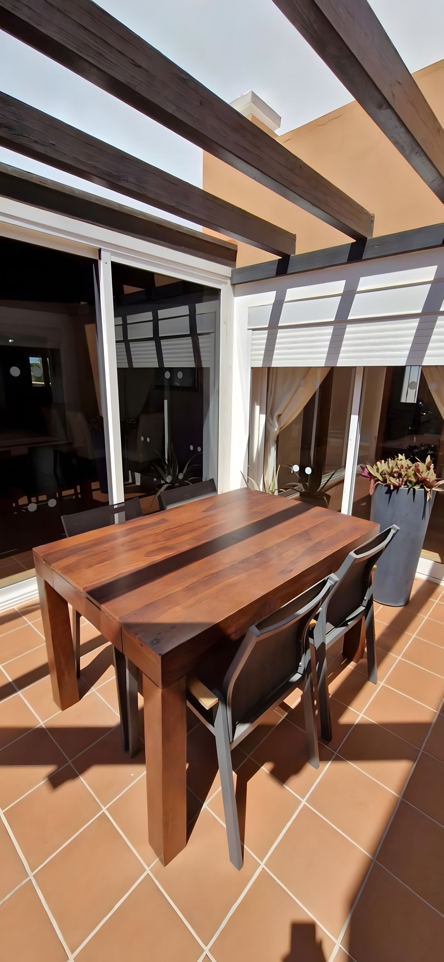 Une terrasse extérieure avec une table et des chaises en bois, un pot de fleurs et une porte-fenêtre coulissante.