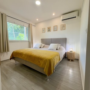 A bedroom with a bed, a window with curtains, an air conditioner, and a bedside lamp.