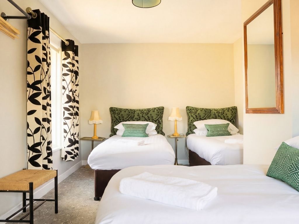 Ett hotellrum med två sängar, en bänk, en spegel och ett fönster med mönstrade gardiner.