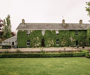 A two-story building with a green ivy-covered exterior, a visible door, and a small garden area with a hedge in the foreground. - Fitz Of Inch - Guest House Rooms (Stradbally)