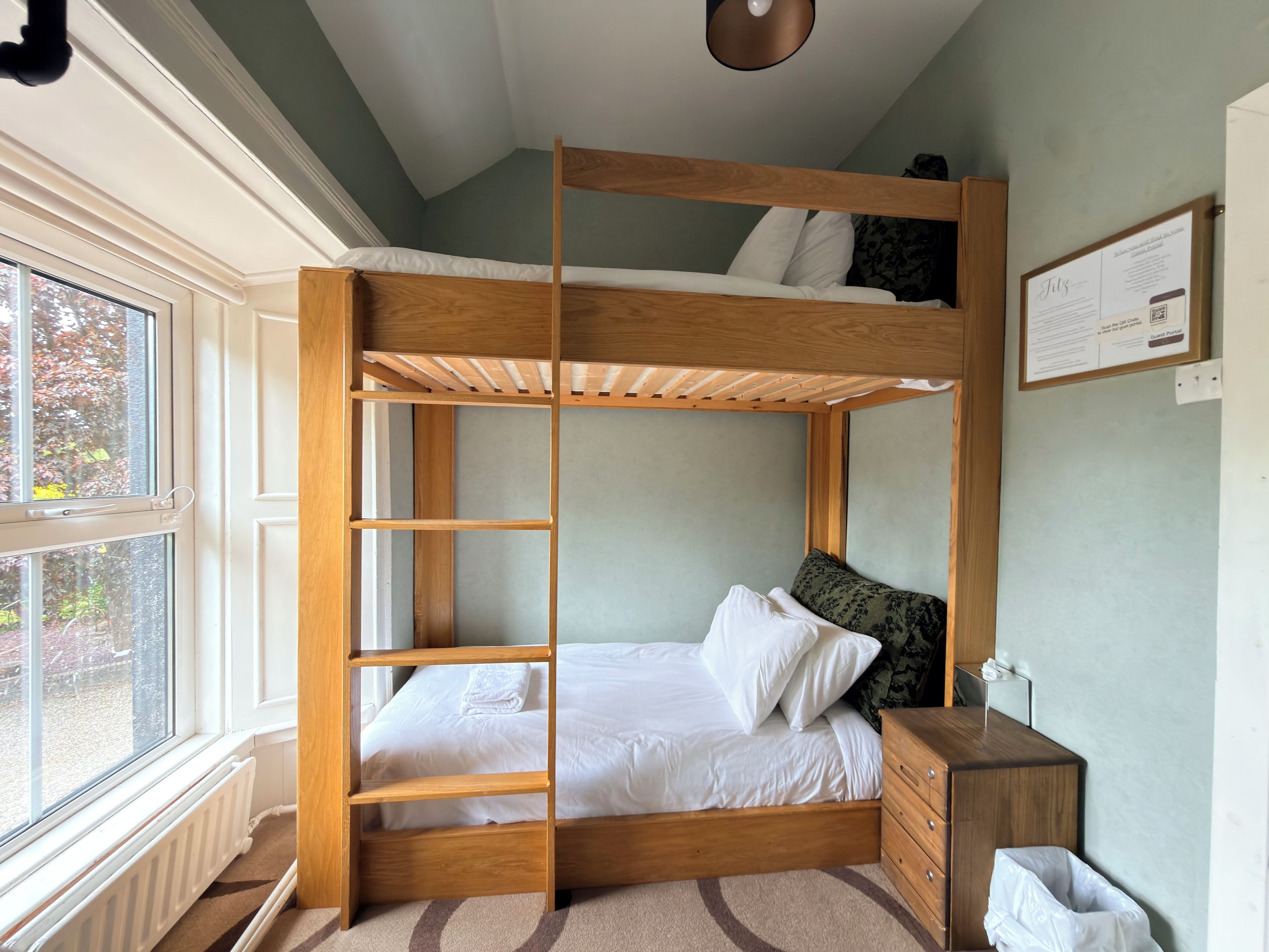 A bunk bed with a wooden frame, a bedside table, a radiator, and a window with a view of trees.