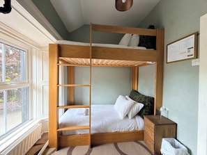 A bunk bed with a wooden frame, a bedside table, a radiator, and a window with a view of trees.