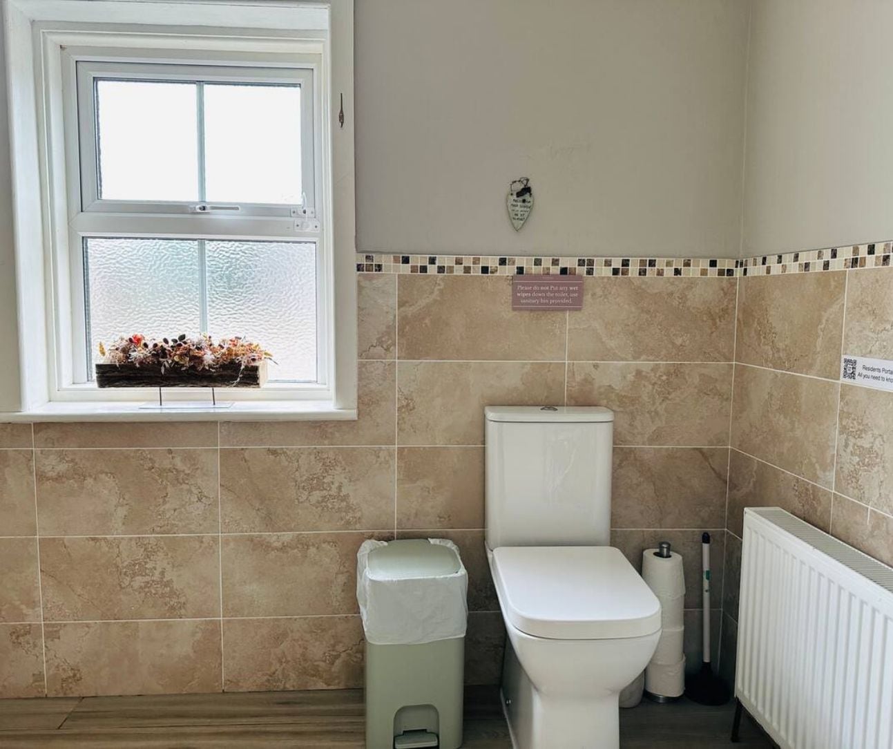 A bathroom with a toilet, a trash bin, a radiator, and a window with a flower arrangement.