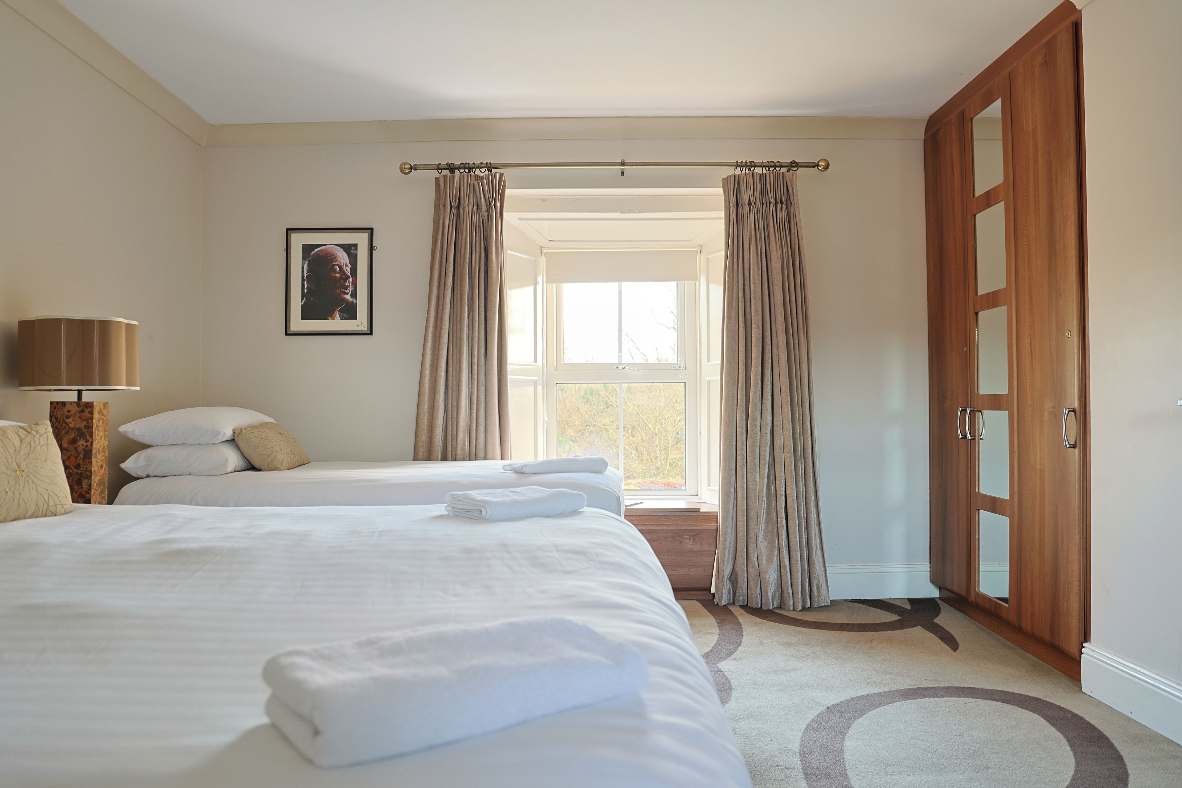 Habitación de hotel con una cama grande, una mesita de noche con lámpara, una ventana con cortinas y una puerta.