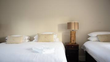 Habitación de hotel con dos camas, cada una con ropa de cama blanca y almohadones decorativos. Un mesita de noche de madera con una lámpara se ubica entre las camas.