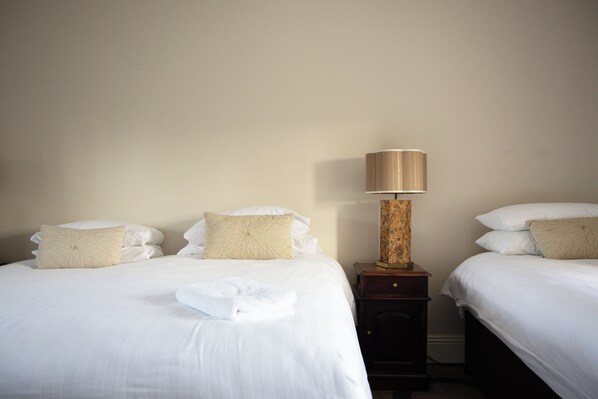 Habitación de hotel con dos camas, cada una con ropa de cama blanca y almohadones decorativos. Un mesita de noche de madera con una lámpara se ubica entre las camas.