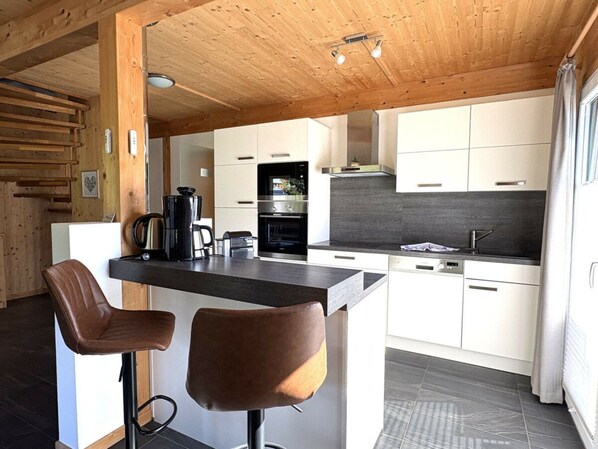 A modern kitchen with a central island, two bar stools, and a built-in oven. - One-bedroom Cabin (Sankt Georgen am Kreischberg)
