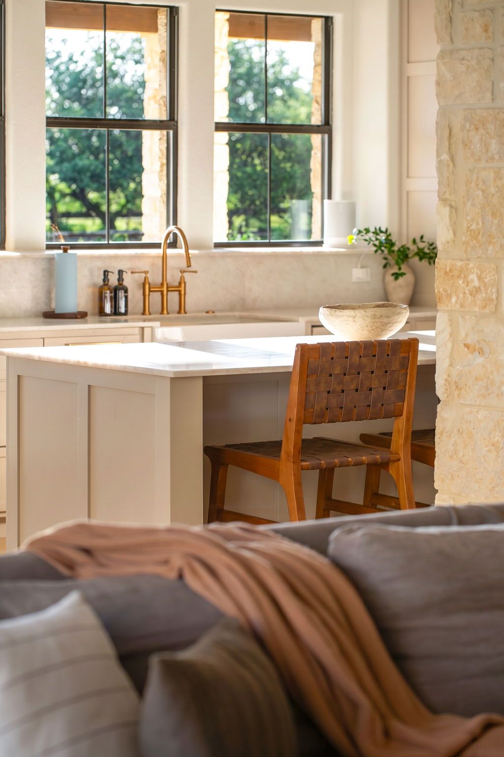 Una cocina con pared de piedra, una silla de madera y vistas al verdor por las ventanas.