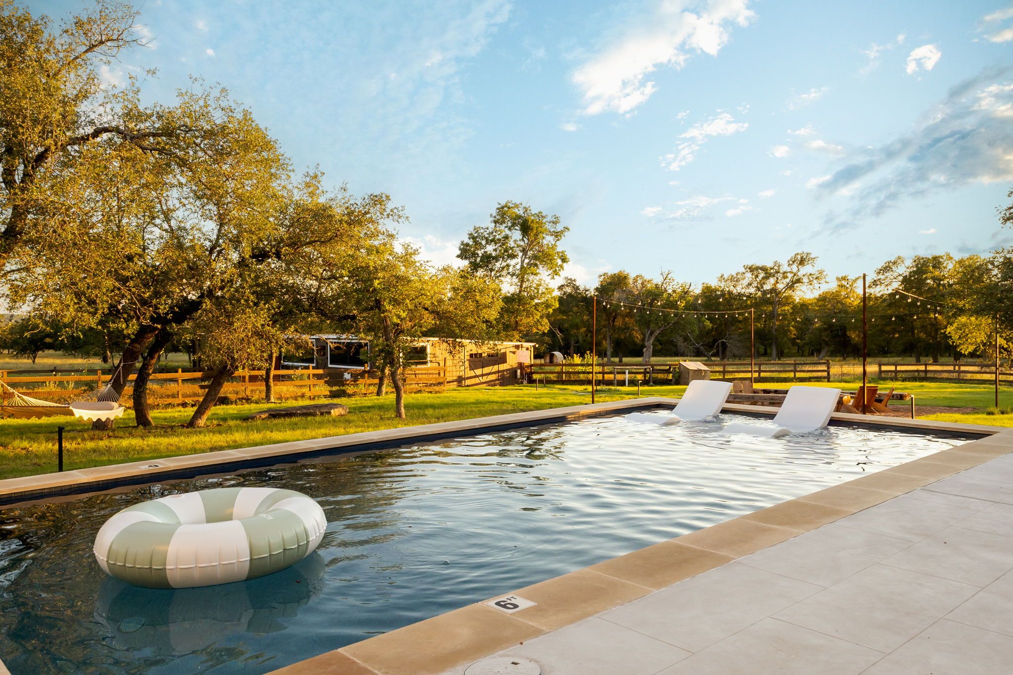 Una piscina con sillas de descanso, un flotador y un área cercada con árboles.