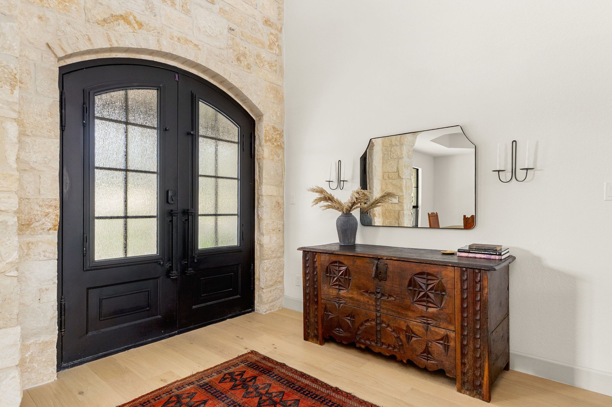 Una habitación con una puerta doble negra, un mueble de madera con adornos tallados, un espejo y un jarrón con plantas secas.