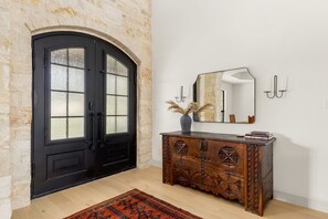 A room with a black double door, a wooden cabinet with carvings, a mirror, and a vase with dried plants.