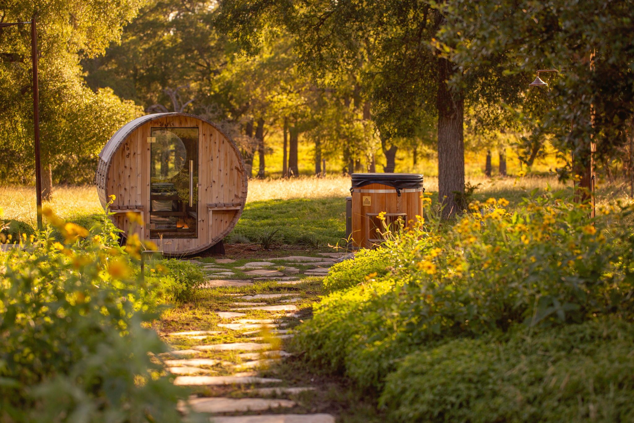 A wooden barrel structure with a glass door, a small wooden shed, and a stone pathway in a forested area.