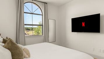Ein Schlafzimmer mit Bett, einem Fenster mit Vorhängen, einem an der Wand befestigten Flachbildfernseher und Blick auf Grünfläche.