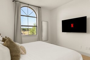 A bedroom with a bed, a window with curtains, a flat-screen TV mounted on the wall, and a view of greenery outside.