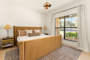 A bedroom with a large bed, two bedside tables, a window with curtains, and a hanging light fixture.
