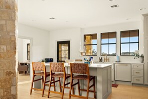 A modern kitchen with a central island, bar stools, and a view of the outdoors through large windows. - Deep Eddy Ranch (Dripping Springs)