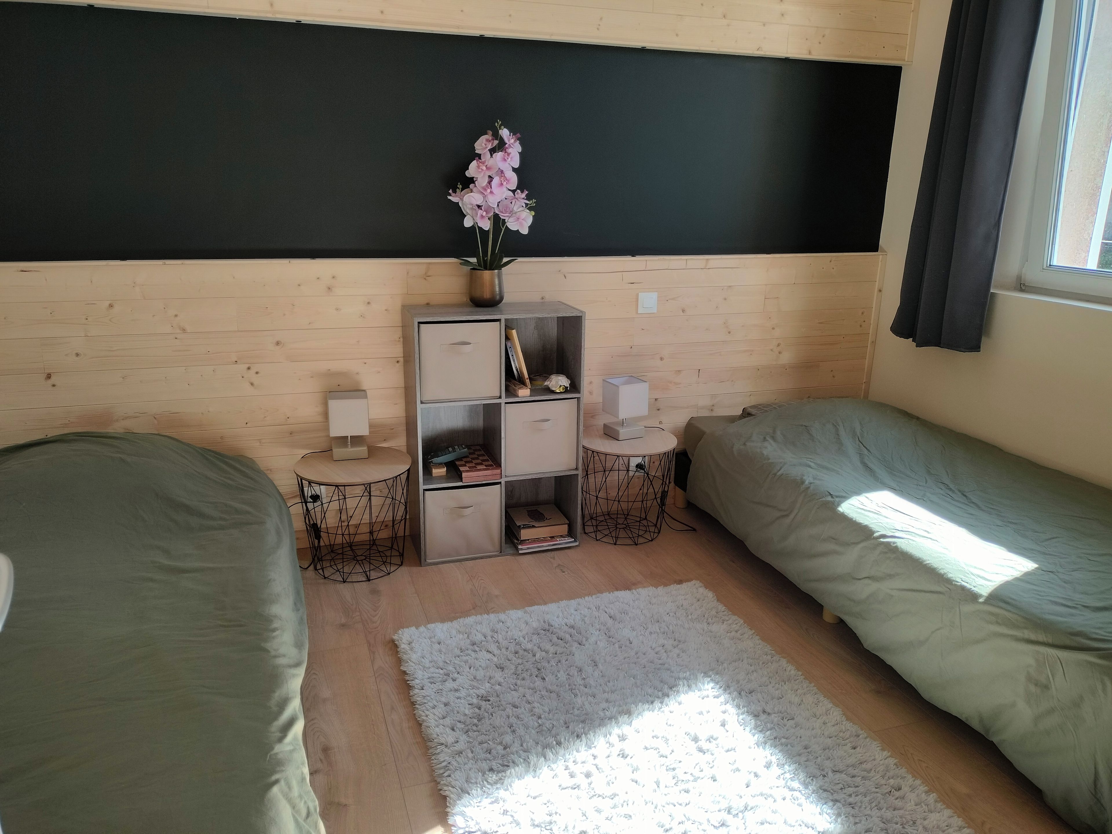 Een slaapkamer met twee bedden, een klein tafeltje met een lamp, een boekenkast en een raam met gordijnen.