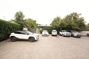 A parking area with several cars, a building with a tiled roof, and a hedge in the background.
