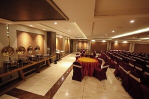 A banquet hall with a long table and rows of chairs, a buffet area with food service equipment, and a wall with decorative plates.