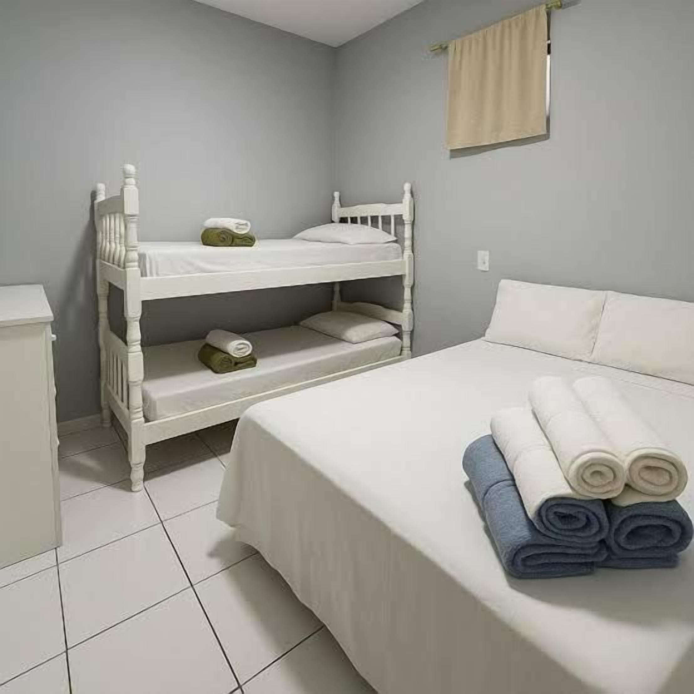 A room with a bunk bed, a single bed, a bedside table, and a rolled-up towel on the bed.