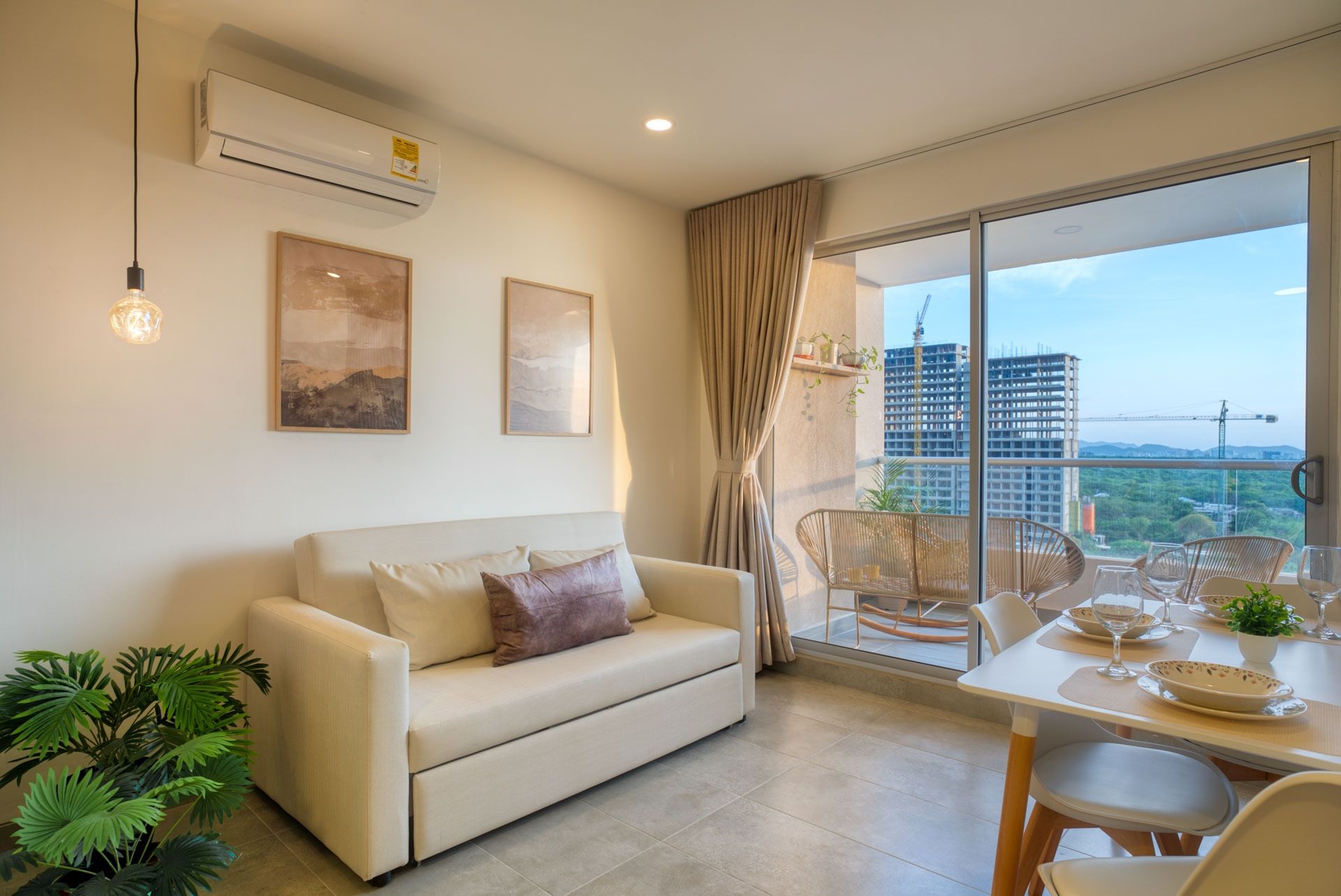 Una sala de estar moderna con sofá, mesa de comedor y sillas, un ventanal con vistas a edificios y un equipo de aire acondicionado en la pared.