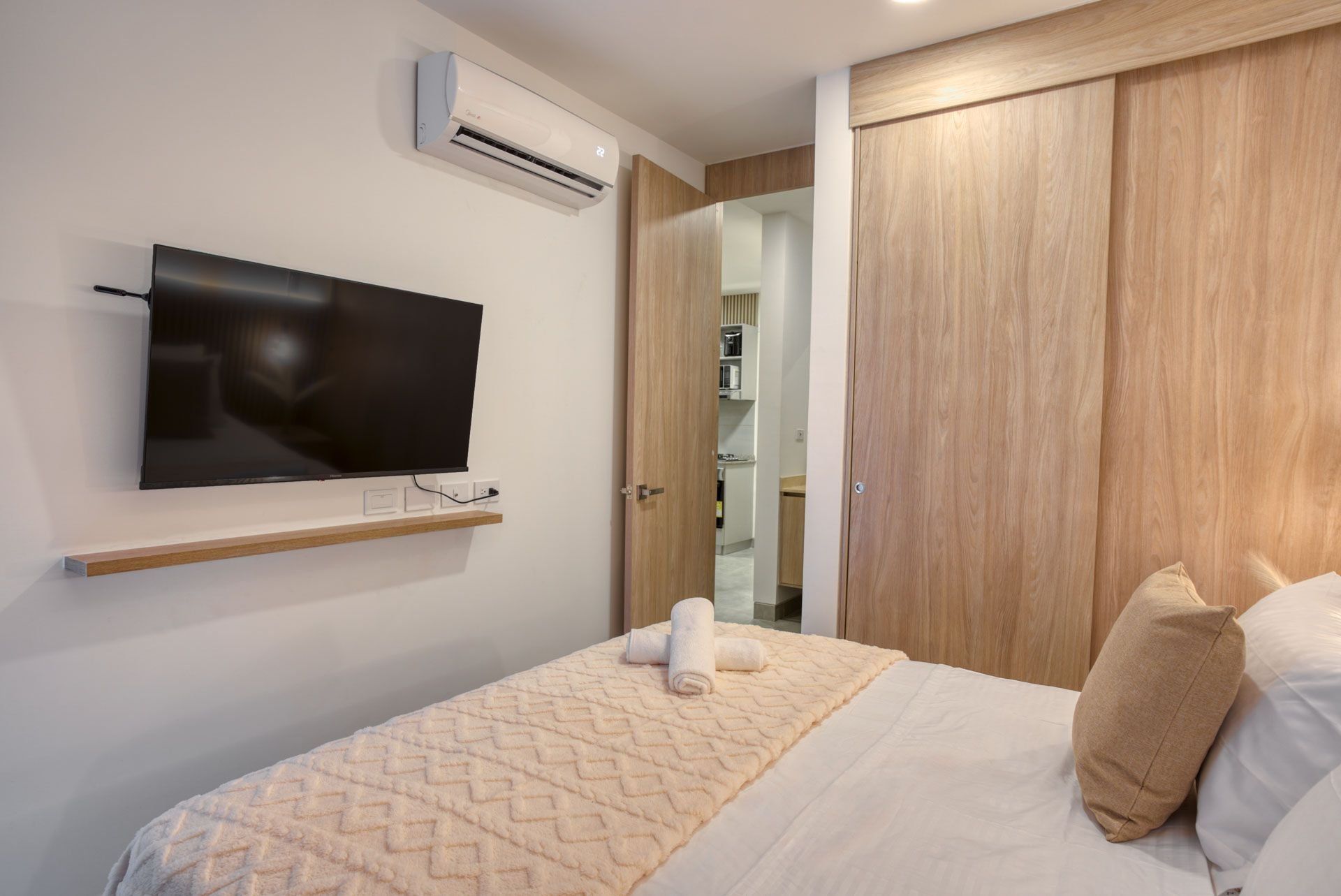 Une chambre à coucher avec un téléviseur à écran plat fixé au mur, une armoire en bois et un lit recouvert d’une couverture à motifs et agrémenté d’oreillers.
