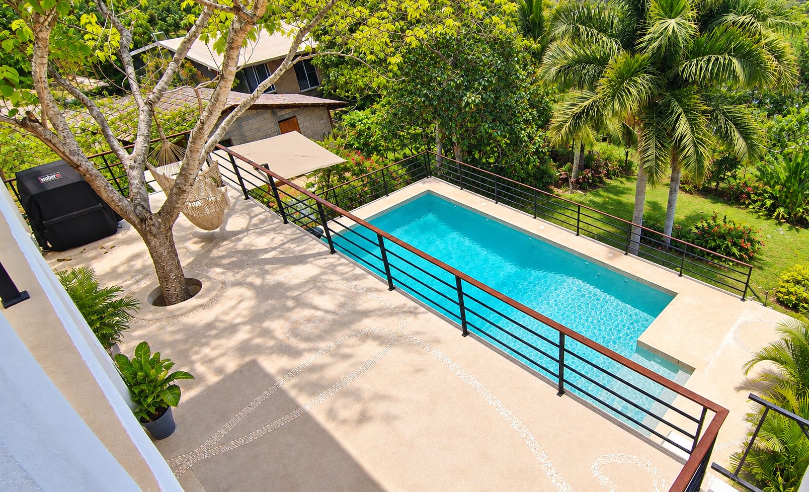 Ein rechteckiger Swimmingpool mit Metallgeländer, umgeben von einer Betonplattform und üppiger Bepflanzung, darunter Palmen und eine Hängematte.