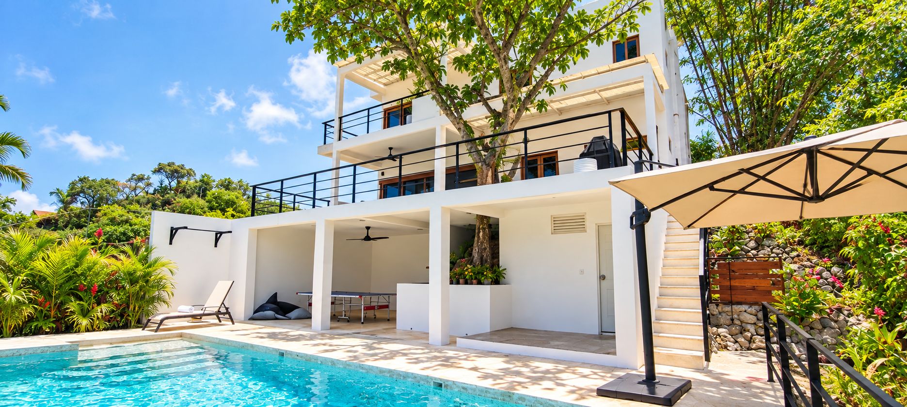 A modern two-story building with a swimming pool, outdoor seating, and lush greenery.