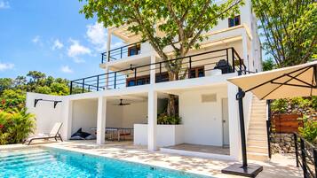 A modern two-story building with a swimming pool, outdoor seating, and lush greenery.