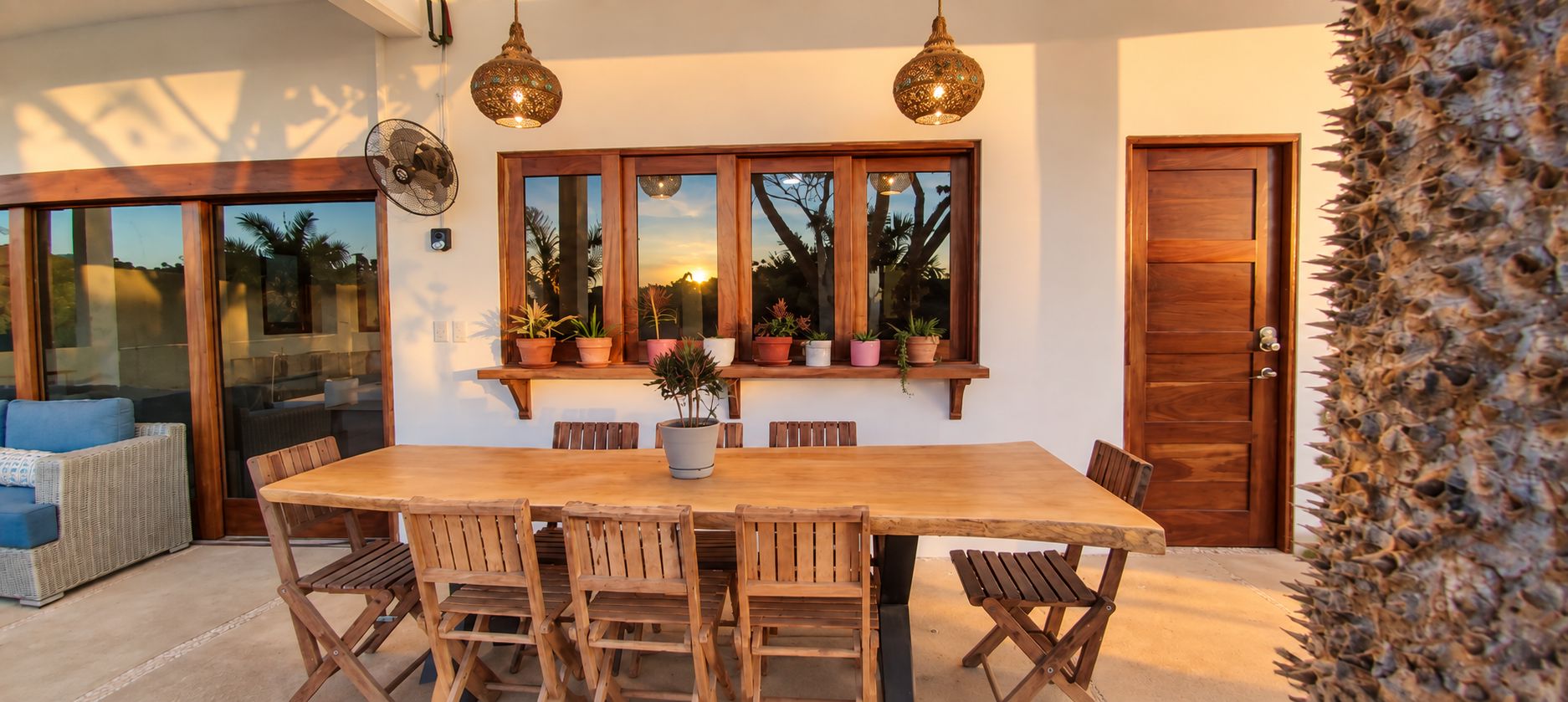 A dining area with a wooden table and chairs, a window with plants, and a door.